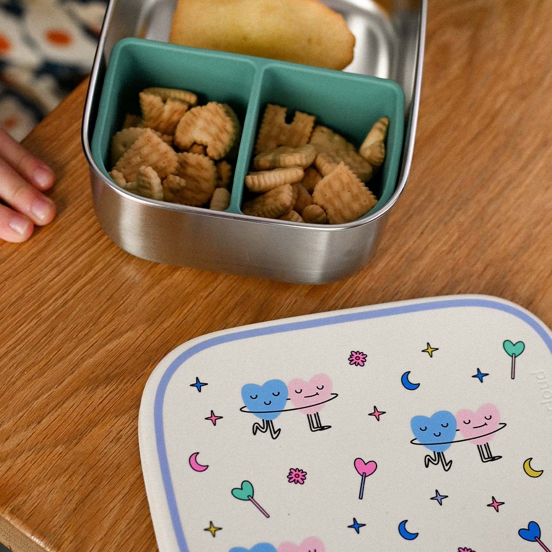 Detail van de the cotton cloud rvs broodtrommel voor kinderen, gevuld met snacks, met het deksel met dancing hearts, sterren en manen patroon op de voorgrond.