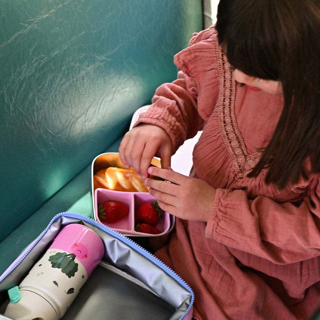 Een kind pakt aardbeien uit de The Cotton Cloud rvs broodtrommel met brood en aardbeien, naast een bijpassende drinkfles in een lunchtas.