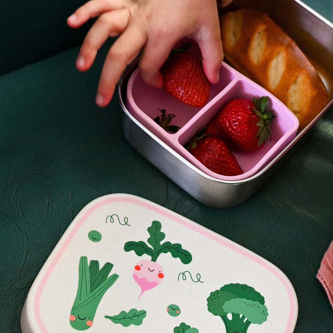 Close-up van een kinderhand die aardbeien pakt uit de The Cotton Cloud rvs broodtrommel met groentefiguren op de deksel op de voorgrond.