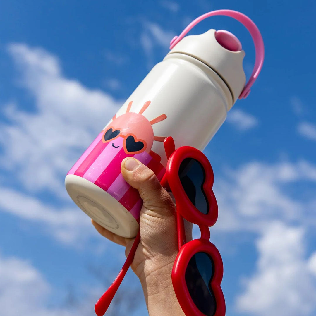 Een hand houdt de The Cotton Cloud rvs drinkfles met zonnetje en roze strepen en een rode hartjeszonnebril omhoog tegen een blauwe lucht.