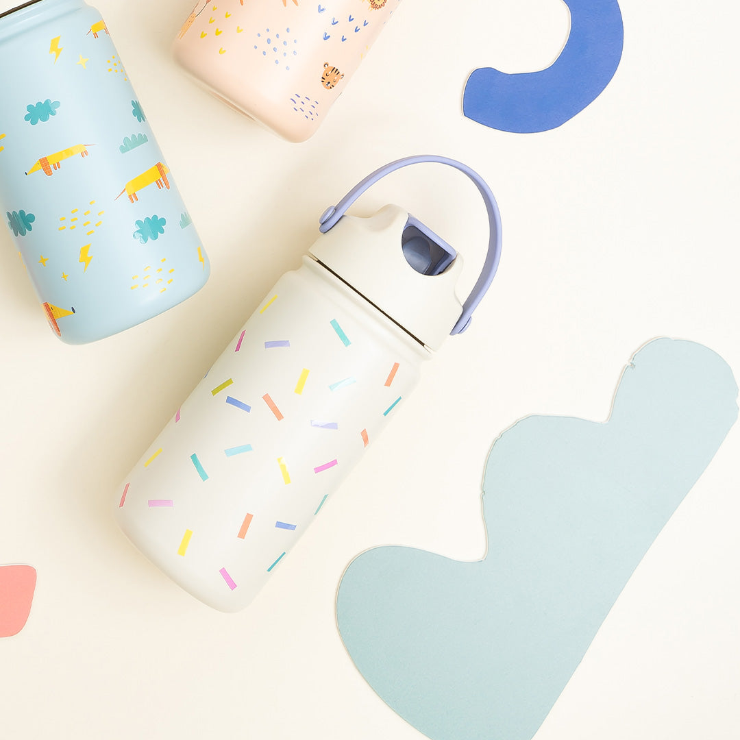 De The Cotton Cloud kinderdrinkfles met confetti patroon ligt tussen andere drinkflessen en papieren decoraties.