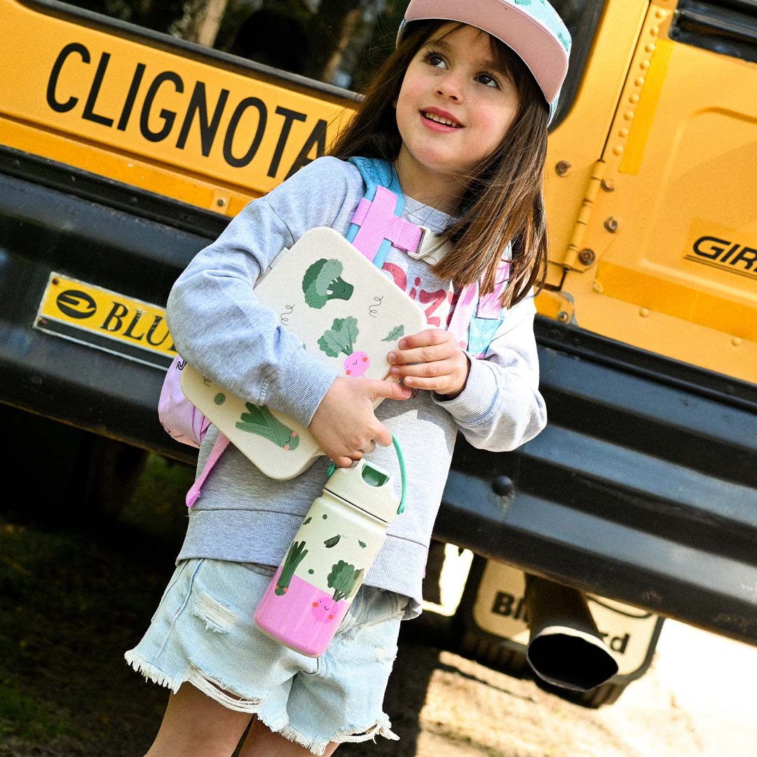 Een meisje staat bij een schoolbus met een kinderdrinkfles en bijpassende lunchbox met groentenfiguren.