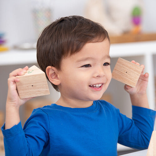 Een lachend kind houdt twee Tickit houten geluiden memorie prismablokken tegen zijn oren.