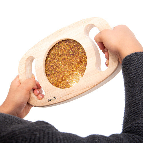 Twee handen houden het houten sensorische glitterpaneel met goudkleurige glitters vast.