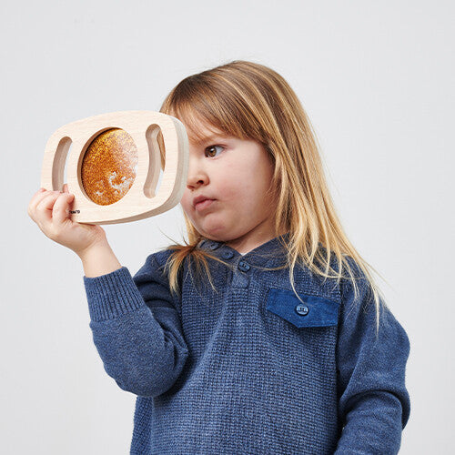 Een jong kind houdt het houten sensorische glitterpaneel met goudkleurige glitters tegen zijn oog.