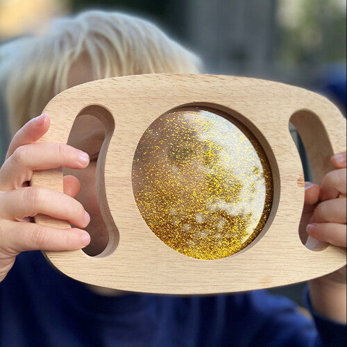 Close-up van handen die het houten sensorische glitterpaneel met bewegende goudkleurige glitters vasthouden.