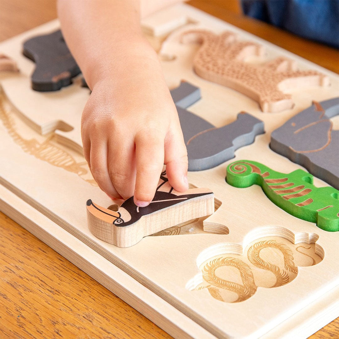 Een kinderhand plaatst een toekan puzzelstukje in een houten safaridieren puzzel.