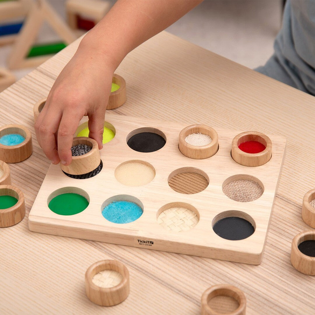 Een kinderhand plaatst een houten cup met grijze textuur in het Tickit sensorisch sorteerspel.