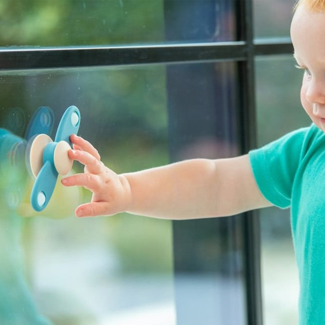 Een baby raakt een blauw-beige tickit silisoft fidget spinner aan die op een raam is bevestigd.