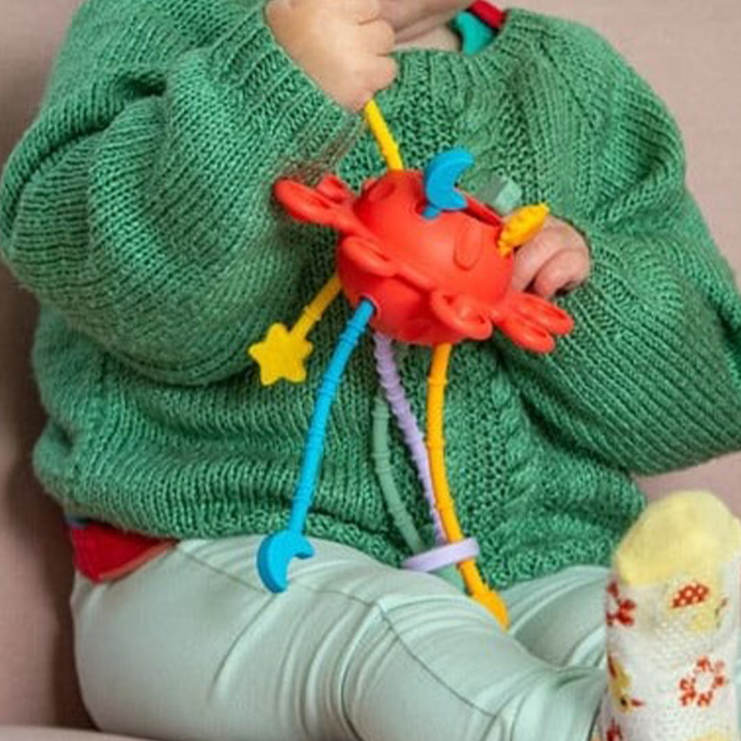 Een baby in een groene trui speelt met het rode tickit silisoft sensorisch activiteitenspeeltje.