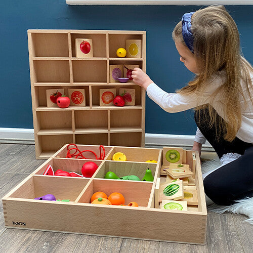 Kind speelt met twee Tickit houten sorteerbakken, één gevuld met houten fruit en groenten, de andere als verticale display.