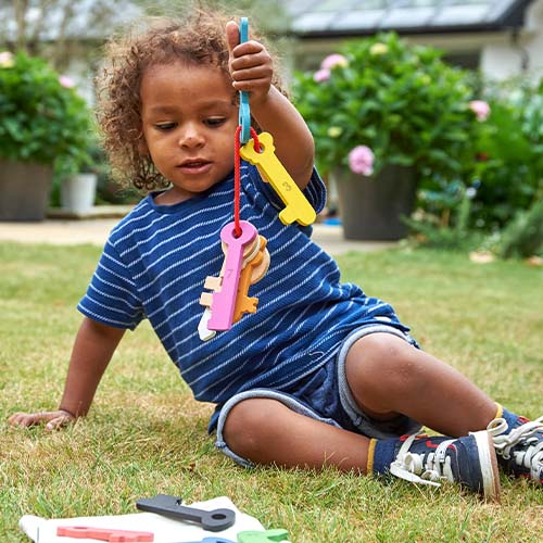 Een kind zit op het gras en houdt een bos kleurrijke Tickit houten babyspeelgoed sleutels omhoog.