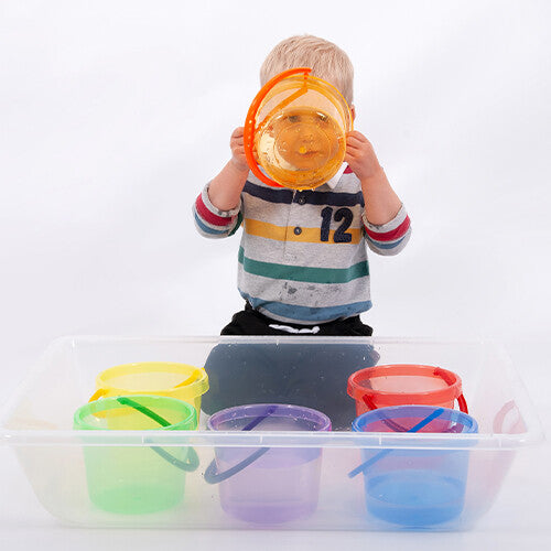 Een kind kijkt door een oranje transparante tickit emmer, met andere emmers in een waterbak.