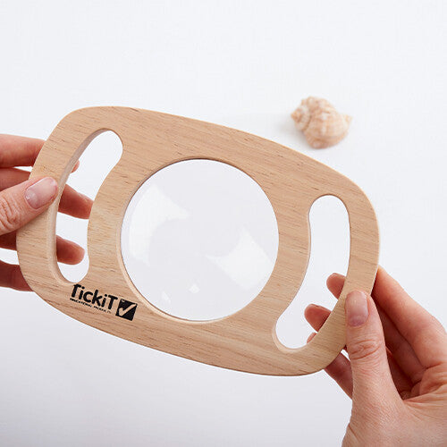 Twee handen houden het Tickit houten vergrootglas vast, met een schelp op de achtergrond.