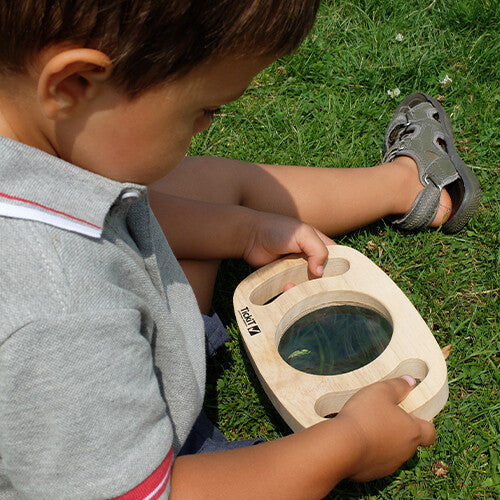 Een kind zit in het gras en kijkt door het Tickit houten vergrootglas.
