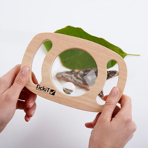 Twee handen houden het Tickit houten vergrootglas vast, dat een vergroot blad en boomschors toont.