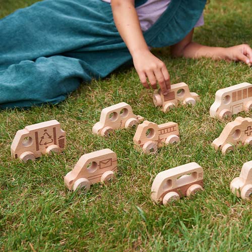 De tickit houten speelgoedvoertuigen liggen verspreid op groen gras, met een kind op de achtergrond.