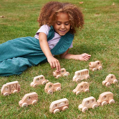 Een lachend kind met krullend haar ligt op groen gras en speelt met de tickit houten speelgoedvoertuigen.