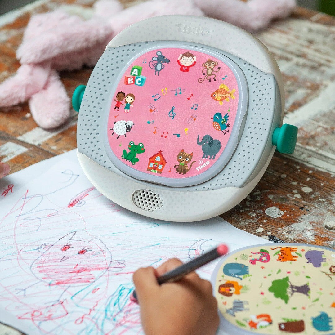 Een kind tekent naast een Timio speler met een roze disc vol dieren, letters en muzieknoten.