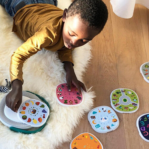 Een kind speelt op een houten vloer met Timio educatieve schijven en een Timio speler.