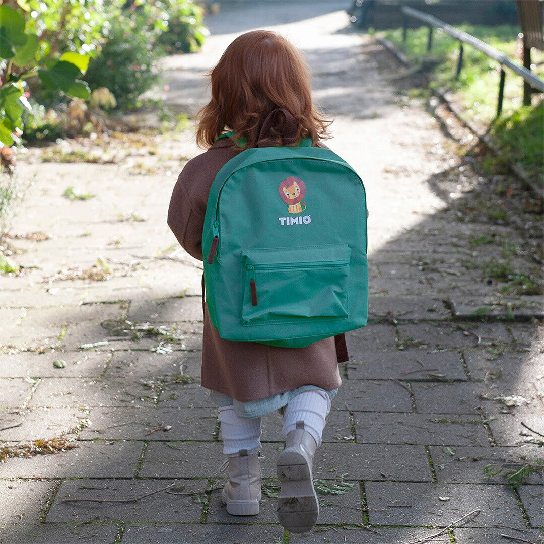 Een kind loopt met de groene Timio kleuterrugzak met leeuwenlogo op de rug over een pad.