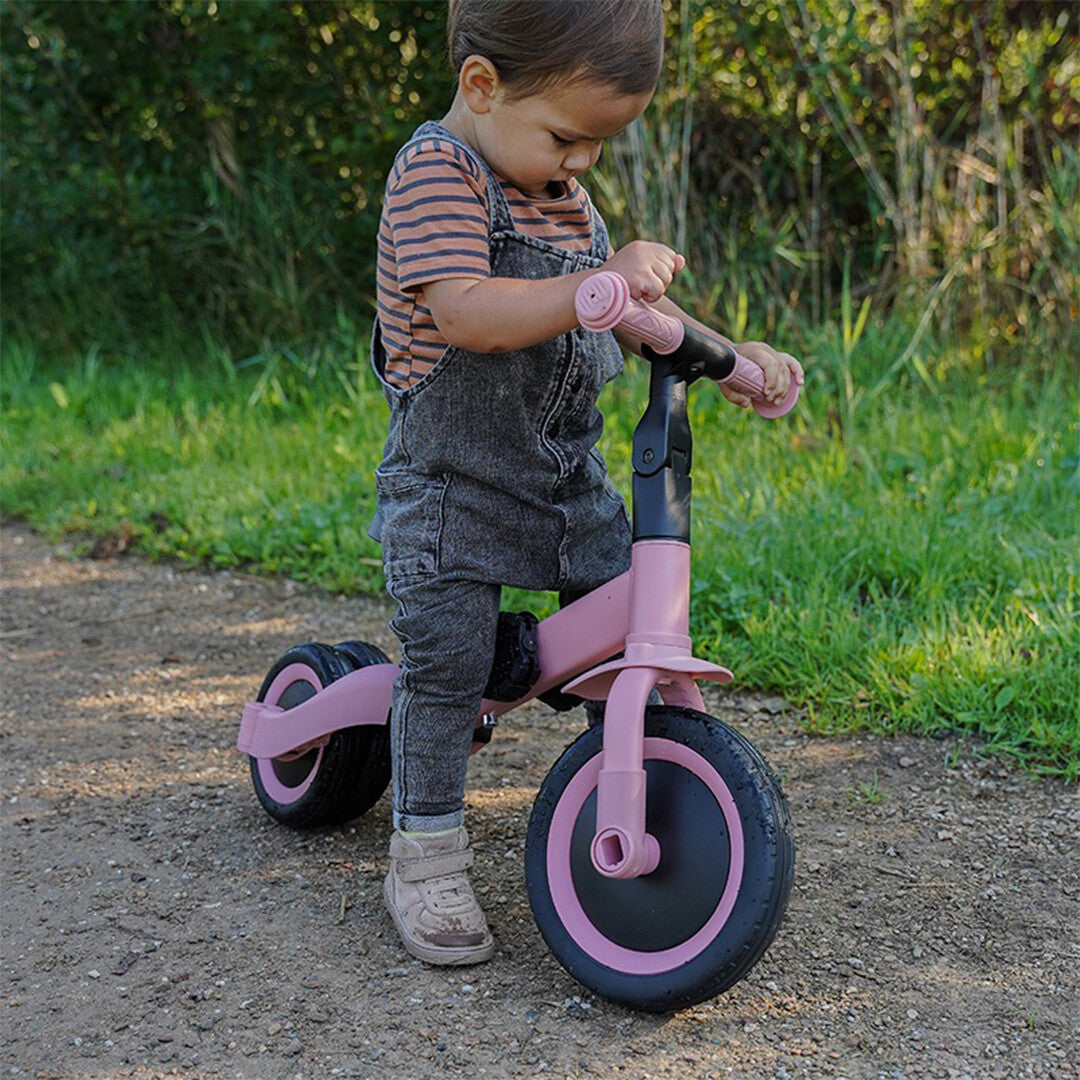 Een jong kind in een tuin staat naast de roze Topmark 4-in-1 loopfiets.