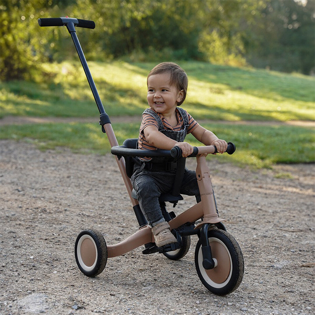 Een lachend kind zit op de Topmark 5-in-1 kinderfiets in driewielerstand met duwstang, buiten op een pad.