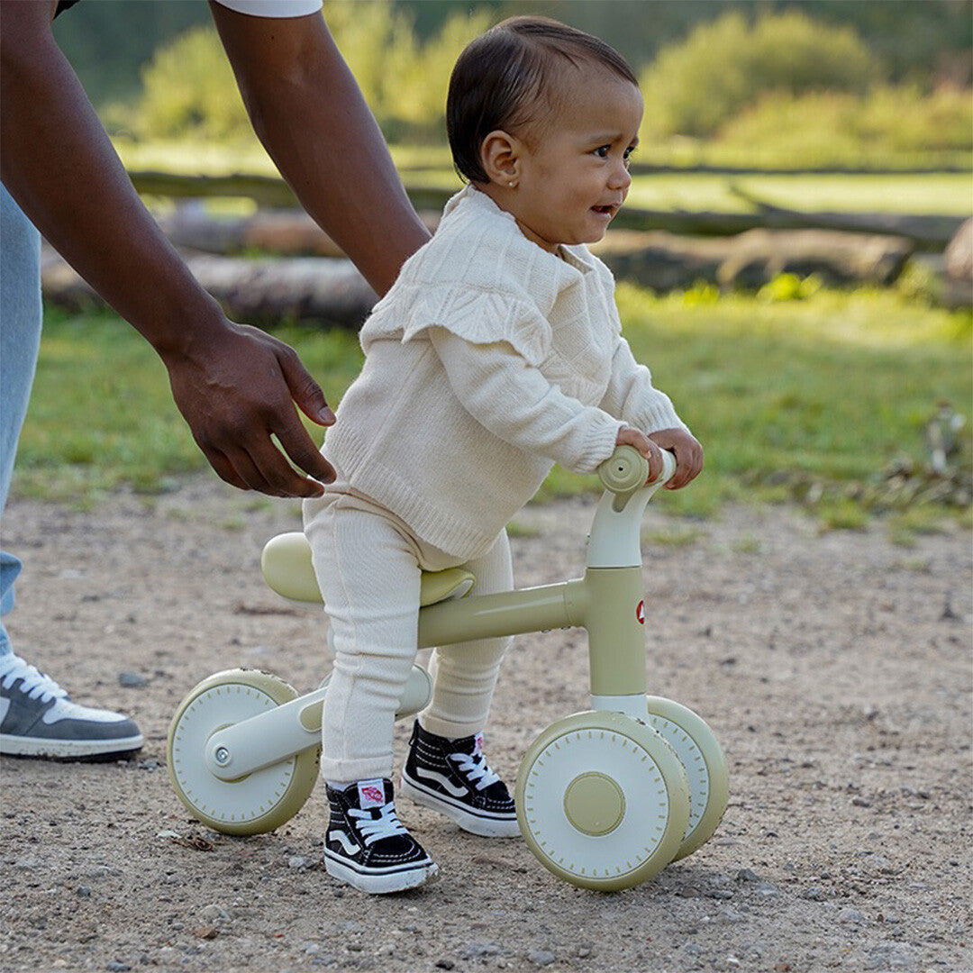 Een baby zit op de Topmark loopfiets Yuki in pistachekleur, ondersteund door een volwassene, buiten op een pad.