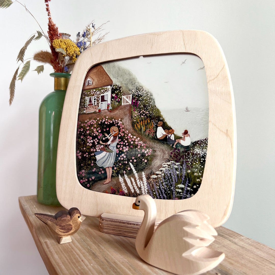 Houten toverplaat lijst met een illustratie van een huis op een klif vol bloemen, staand in een houten standaard.