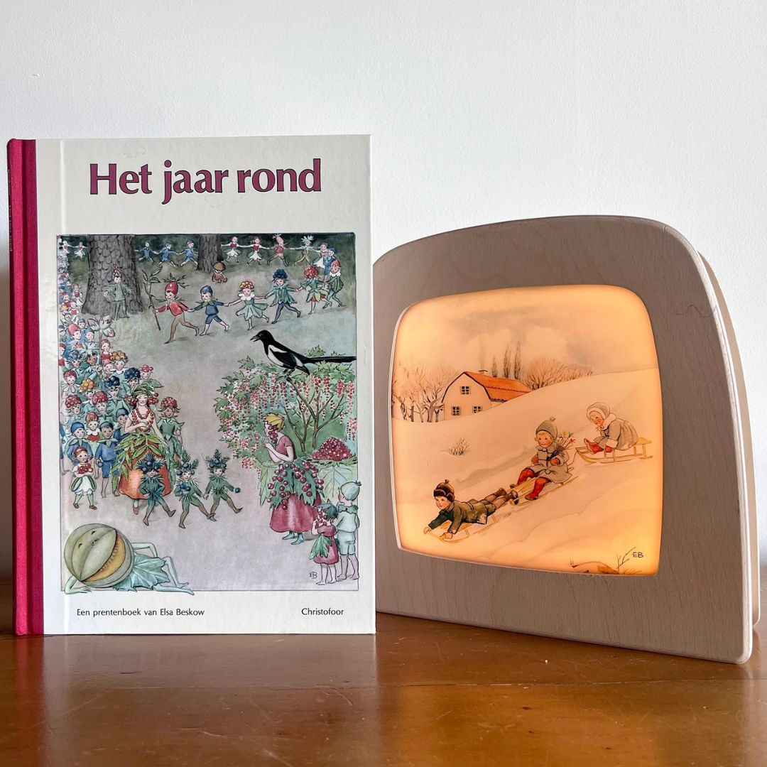 Een Toverlux toverlamp met de toverplaat van sleeënde kinderen naast het prentenboek 'Het jaar rond'.