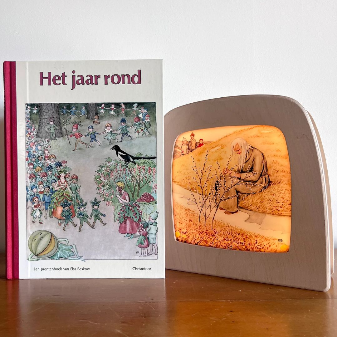 Het boek 'Het jaar rond' naast een Toverlux toverlamp met de illustratie van een oude man en kinderen bij wilgenkatjes.