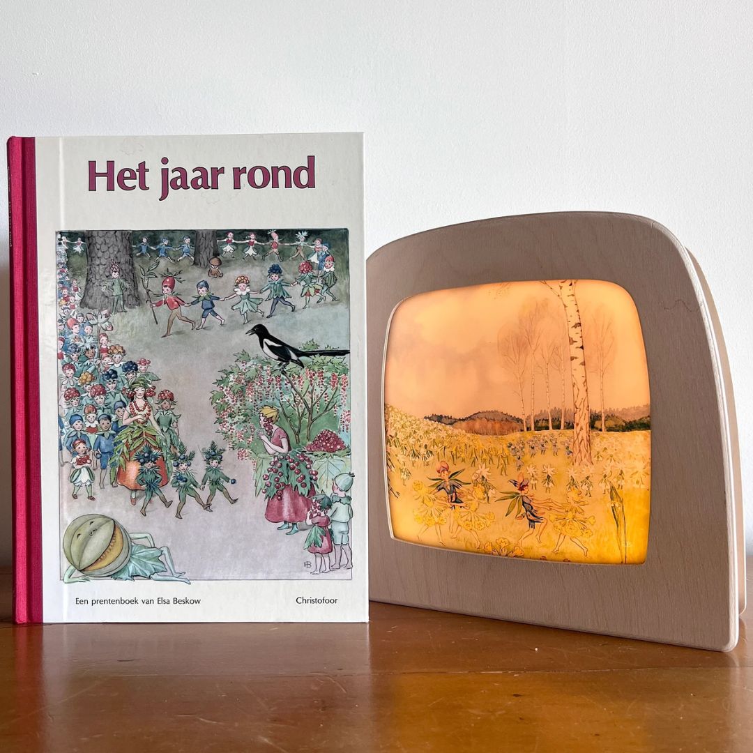 Een Toverlux toverlamp met de 'mei' toverplaat naast een boek getiteld 'Het jaar rond' van Elsa Beskow.