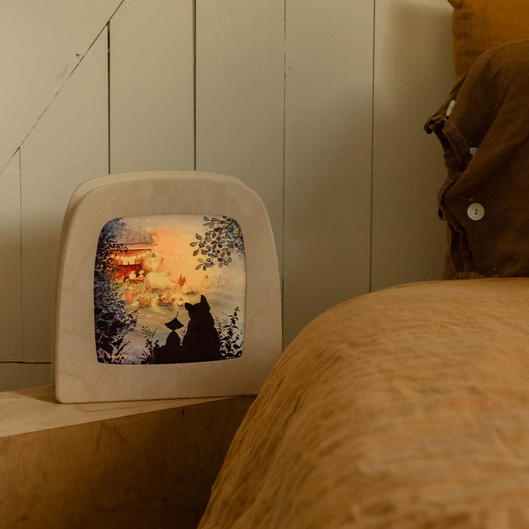 Toverlux toverplaat met schuurfeest-illustratie, verlicht en staand op een nachtkastje naast een bed.
