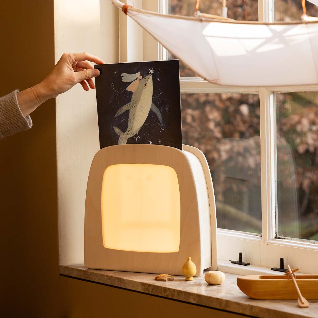 Een hand haalt de Toverlux walvis toverplaat uit de verlichte toverlamp op een vensterbank.