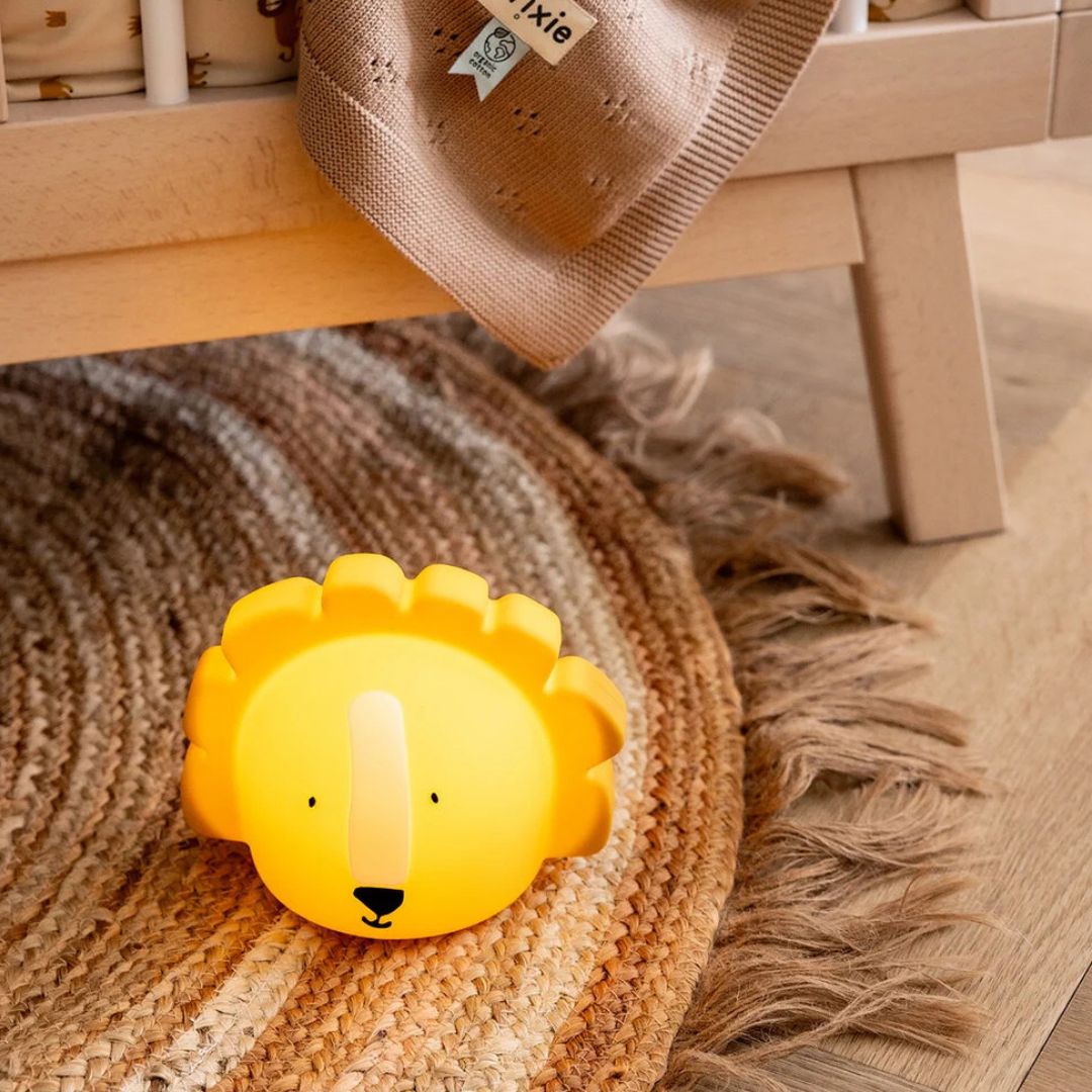 Het Trixie leeuw nachtlampje staat lichtgevend op een gevlochten vloerkleed naast een babybedje.