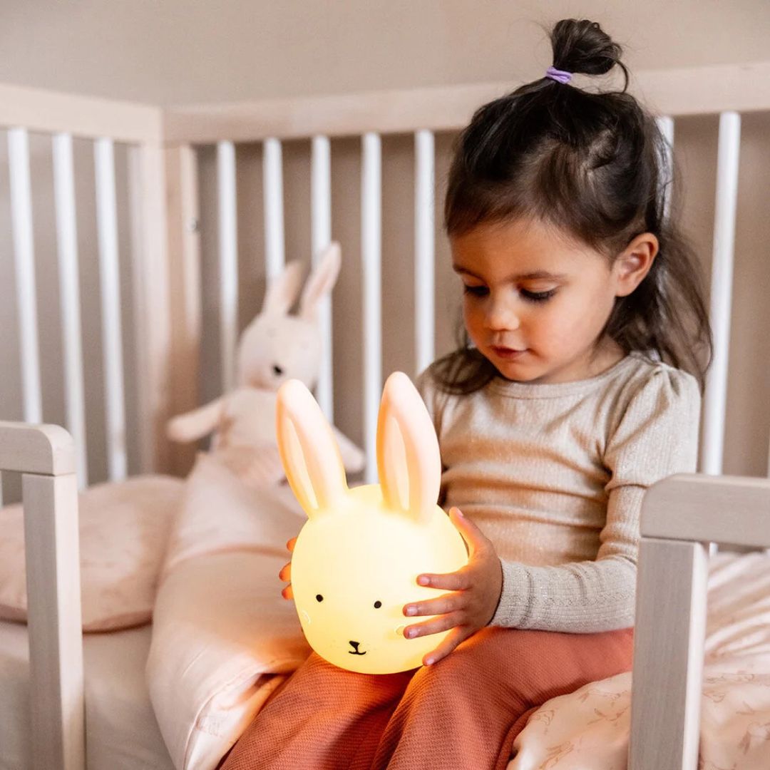 Een meisje zit in een ledikant en houdt het Trixie nachtlampje in de vorm van een lichtgevend konijn vast.