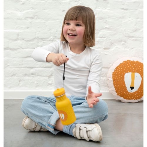 Een lachend kind zit op de grond en houdt de gele Trixie drinkfles met leeuwenkop en zwarte draaglus vast.