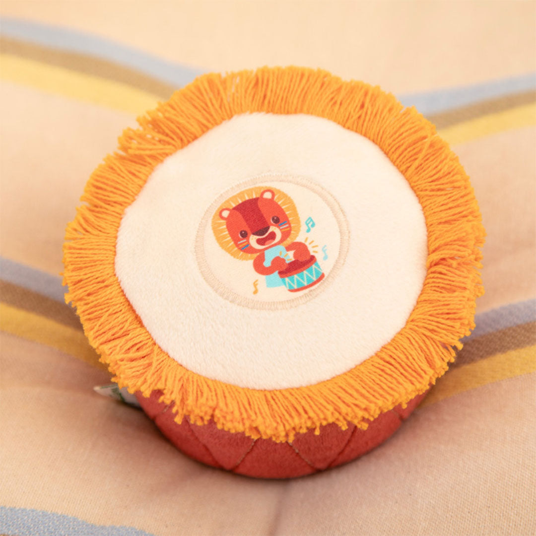 Lilliputiens babyspeelgoed rammelaar in de vorm van een oranje trommel met een leeuw erop, liggend op een gestreepte ondergrond.