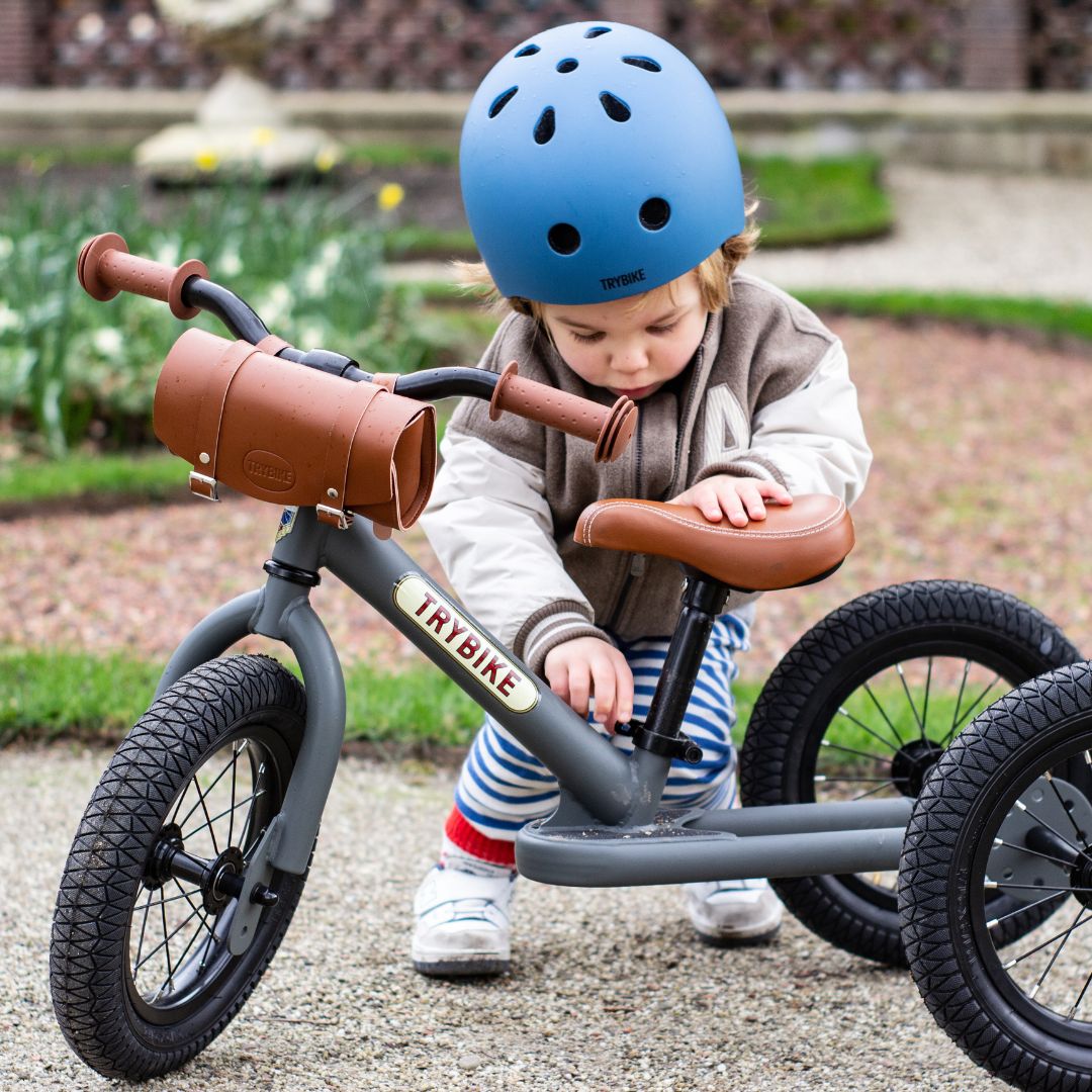 Een kind met blauwe helm stelt een grijze Trybike driewieler met bruine stuurtas buiten af.