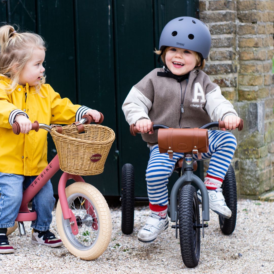 Twee kinderen spelen buiten met een grijze Trybike driewieler en een roze Trybike loopfiets.