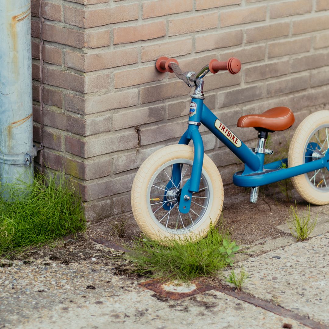 Trybike stalen 2-in-1 loopfiets in vintage blauw als tweewieler, leunend tegen een bakstenen muur buiten.