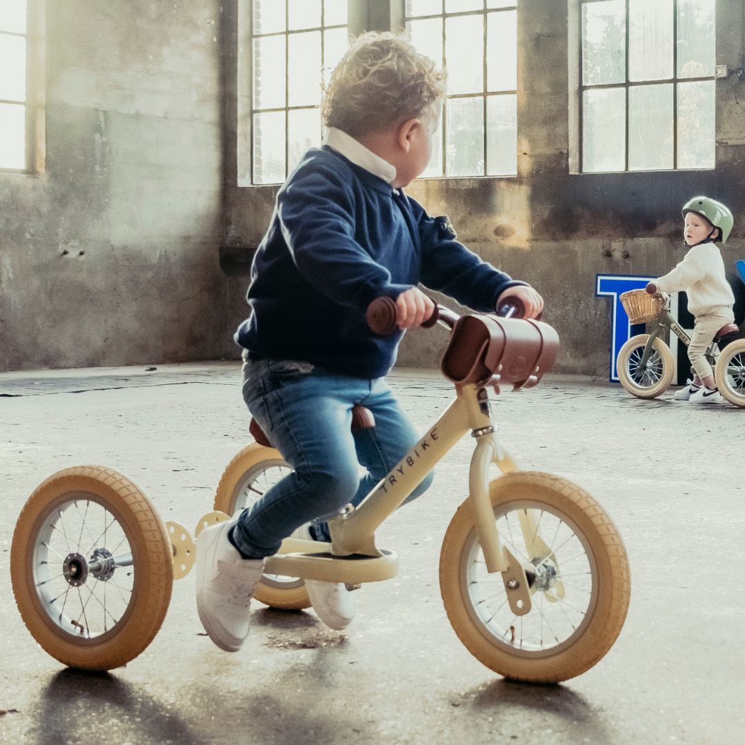 Een kind rijdt op een Trybike 2-in-1 loopfiets als driewieler in vintage cream, met een ander kind op een loopfiets op de achtergrond.