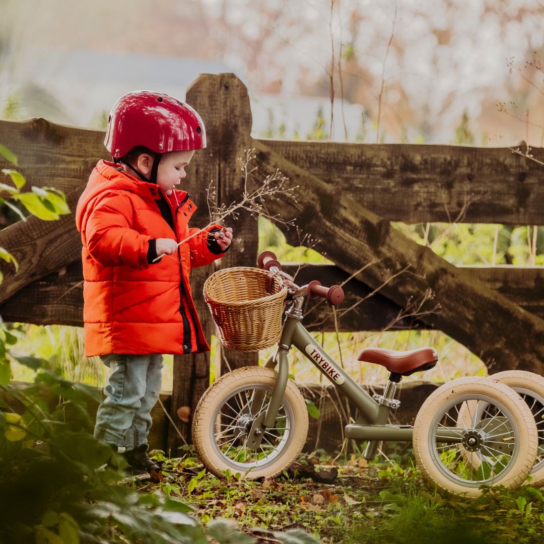 Een kind met rode jas en helm staat naast de Trybike 2-in-1 loopfiets met drie wielen in een bos.