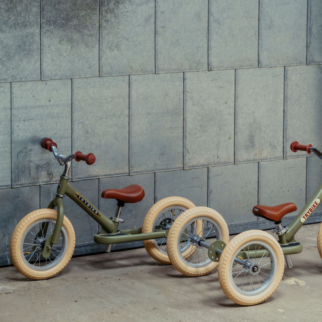 Twee Trybike 2-in-1 loopfietsen, één als driewieler en één als tweewieler, staan tegen een grijze muur.
