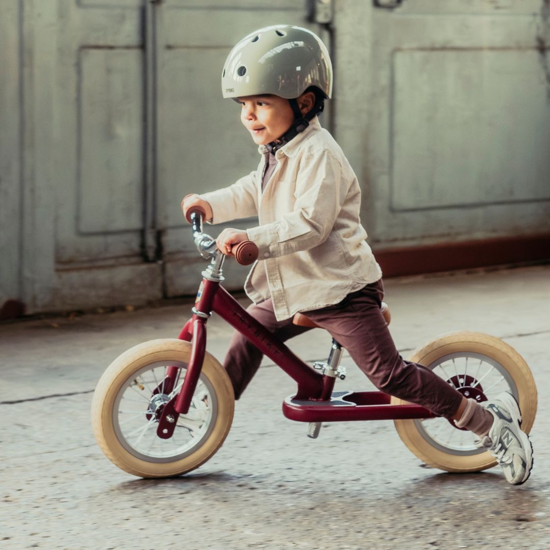 Een jong kind met helm rijdt op de Trybike vintage rode loopfiets als tweewieler.