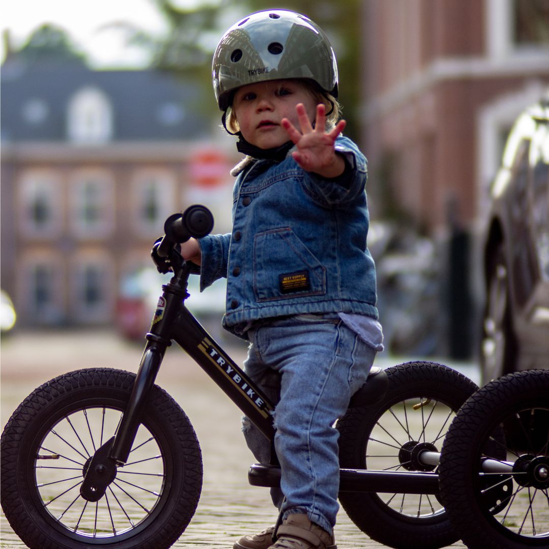 Een kind met helm en spijkerjasje zit op een zwarte Trybike driewieler op een klinkerstraat en zwaait naar de camera.