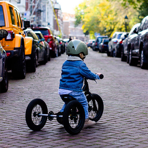 Een kind met helm en spijkerjasje rijdt op een zwarte Trybike driewieler op een klinkerstraat, van achteren gefotografeerd.