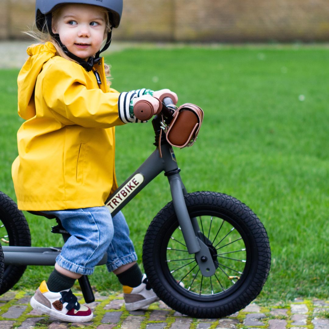 Een kind met gele regenjas en helm zit op een Trybike stalen loopfiets buiten.