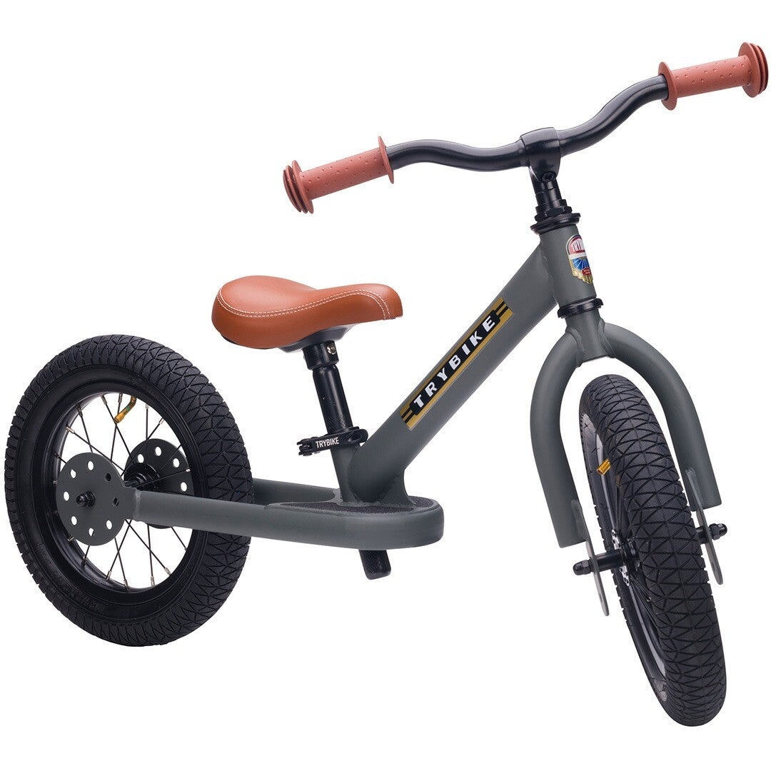 Trybike stalen loopfiets in matgrijs met bruin zadel en handvatten, schuin van voren gezien.