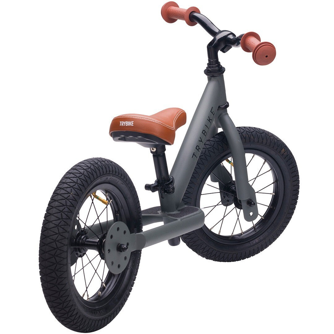 Trybike stalen loopfiets in matgrijs met bruin zadel en handvatten, schuin van achteren gezien.